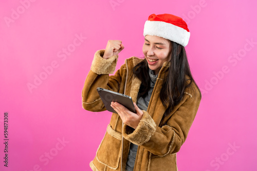 Pretty Asian girl in long sleeve shirt and Christmas hat. holding tablet in hand and expresses delight in ordering  buy online products at a special price. Indoor studio isolated on pink background photo