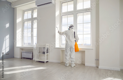 Professional pest control inside the house. Exterminator wearing protective suit, mask and gas respirator spraying insecticide from yellow sprayer bottle over white window frame in modern room at home