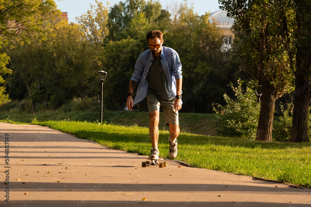 Fototapeta premium Front view of young guy riding on longboard along empty asphalt road between green trees