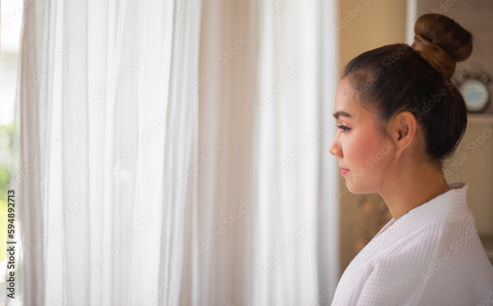 Side view of charming elegant middle aged asian woman in white bathrobe standing to the window in spa salon while looking to the copy space. Side view of charming elegant middle aged asian woman in white bathrobe standing to the window in spa salon while looking to the copy space.