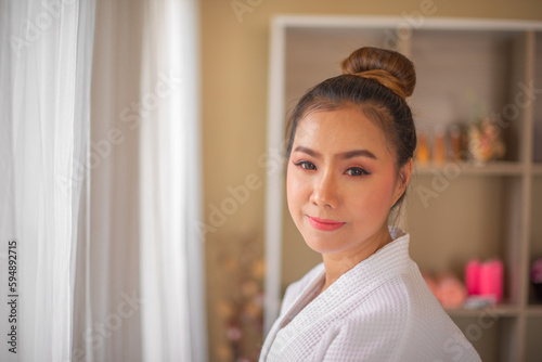 Side view of charming elegant middle aged asian woman in white bathrobe standing with smile enjoying spa procedures in spa salon while looking to the camera.