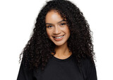 Optimistic curly young woman with gentle smile, looks positively at camera, wears casual black clothes, isolated on grey background, listens interesting story or funny anecdote. Emotions concept