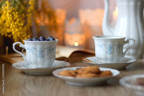 Cup of tea or coffee  plate of cookies  cup of blueberries  plate of chocolate  glass of juice  book  reading glasses  teapot  flowers and lit candles on the table. Brunch or afternoon tea concept.