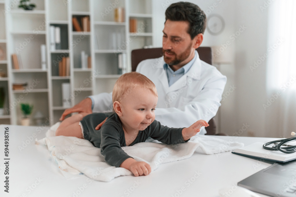 Toddler is lying down on the table. Doctor with little baby is working in the cabinet Toddler is lying down on the table. Doctor with little baby is working in the cabinet