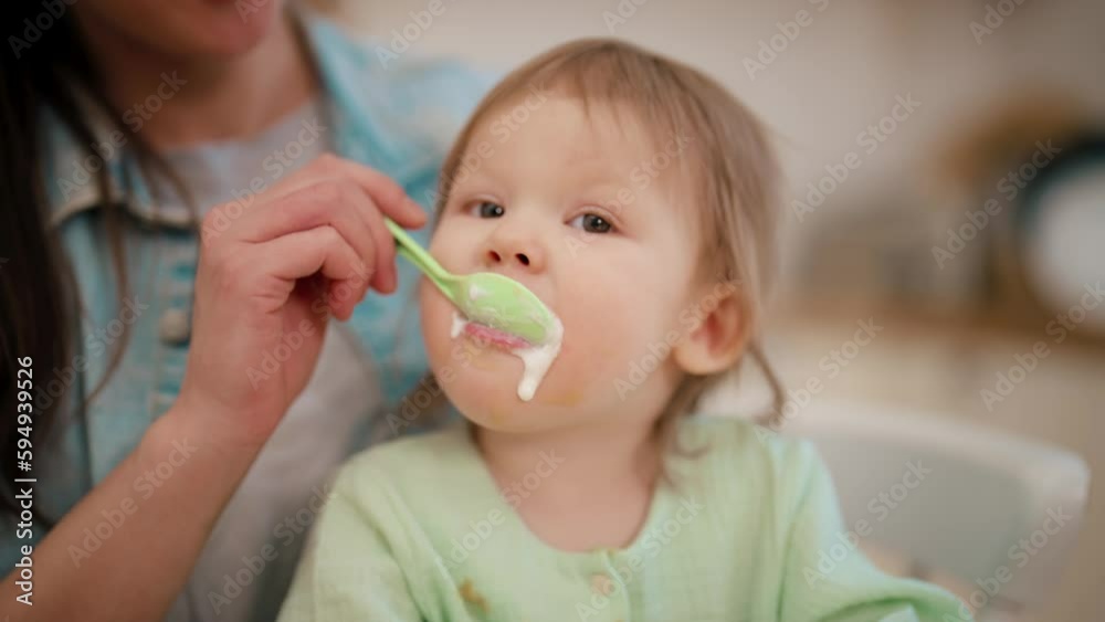 Mom feeds her baby playing with food. Toddler eats fruit or vegetable puree. Mother holds child on knees at table on kitchen. Raising child, kid's nutrition, motherhood, love care in family concept. Mom feeds her baby playing with food. Toddler eats fruit or vegetable puree. Mother holds child on knees at table on kitchen. Raising child, kid's nutrition, motherhood, love care in family concept.