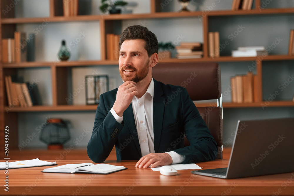 Sitting by table with laptop. Businessman in formal clothes is working indoors Sitting by table with laptop. Businessman in formal clothes is working indoors