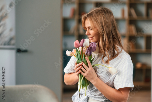 Positive young woman is standing in domestic room with flowers