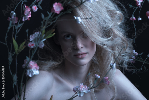 Beautiful woman with big wavy hair and purple make-up studio portrait. Model surrounded with flower twigs with blossoms with naked shoulders looking at camera. Toned image with blue color