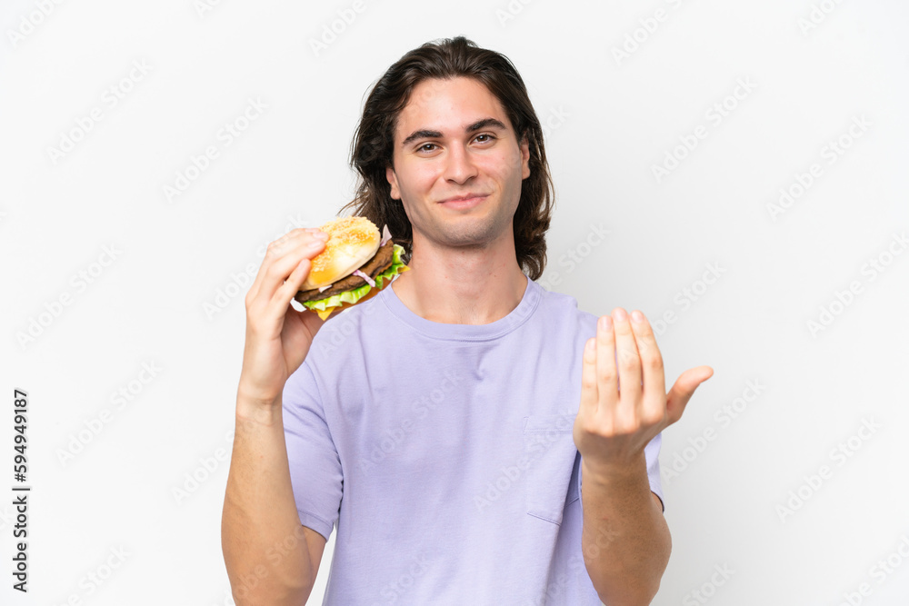 Young handsome man holding a burger isolated on white background inviting to come with hand. Happy that you came Young handsome man holding a burger isolated on white background inviting to come with hand. Happy that you came