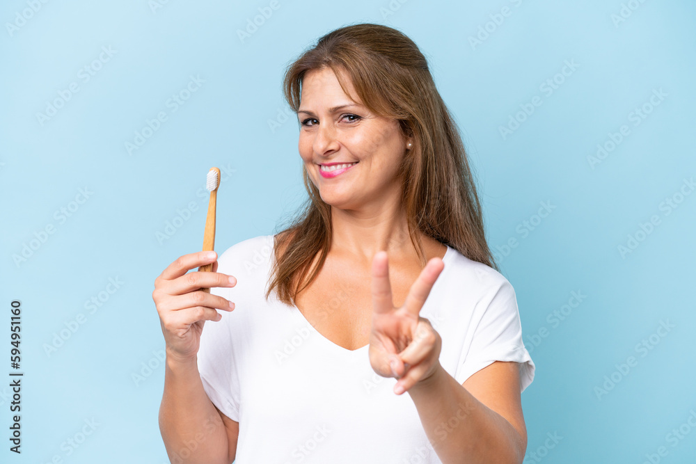 Middle-aged caucasian woman brushing teeth isolated on blue background smiling and showing victory sign Middle-aged caucasian woman brushing teeth isolated on blue background smiling and showing victory sign