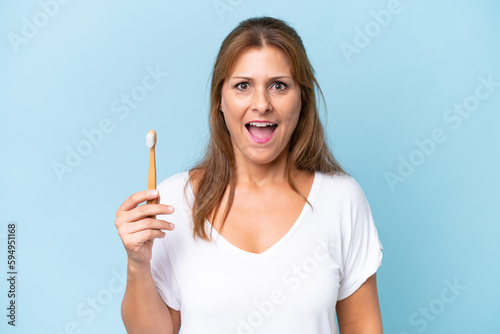 Middle-aged caucasian woman brushing teeth isolated on blue background with surprise and shocked facial expression