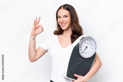 Young caucasian woman isolated on white background holding a weighing machine and doing OK sign