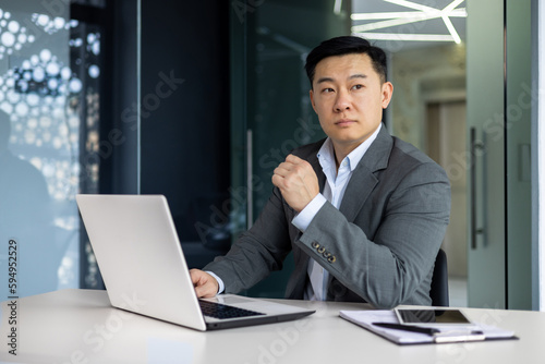 Serious thinking asian businessman working inside office sitting at table with laptop  boss making important financial decision  asian mature man in business suit at workplace.