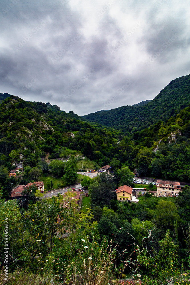 Small town in the middle of the Asturias mountain, with green grass, large mountains in the background and cloudy sky.
