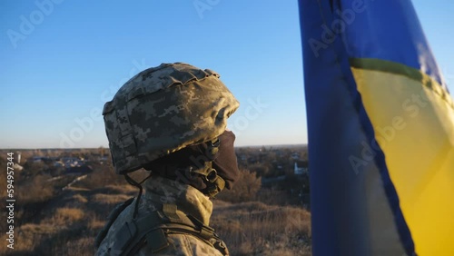 Dolly shot of male soldier in military uniform and helmet holds a waving flag of Ukraine to peak of hill. Ukrainian army man stands with lifted national banner against blue sky photo