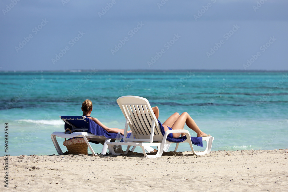 Two girls in bikini tanning on white deck chairs on sea waves background. Vacation on sandy beach