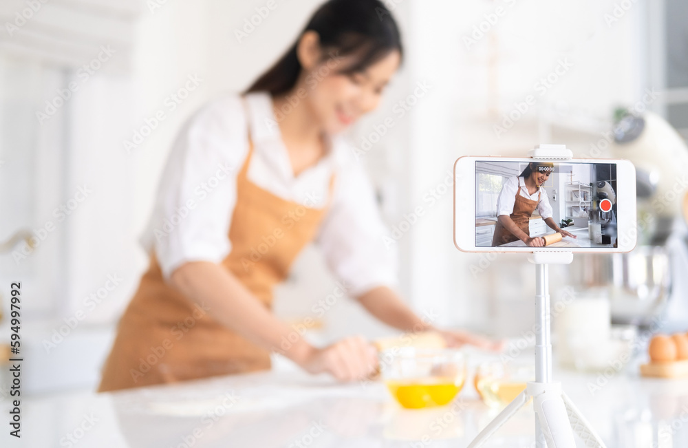 baker online courses. food preparing and culinary training class concept. smiling asian chef kneading dough in the kitchen and shooting video of herself using mobile phone on a tripod. baker online courses. food preparing and culinary training class concept. smiling asian chef kneading dough in the kitchen and shooting video of herself using mobile phone on a tripod.