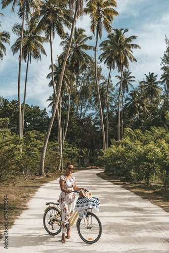 Pretty woman on bicycle on sandy path surrounded with tall palm trees on tropical island