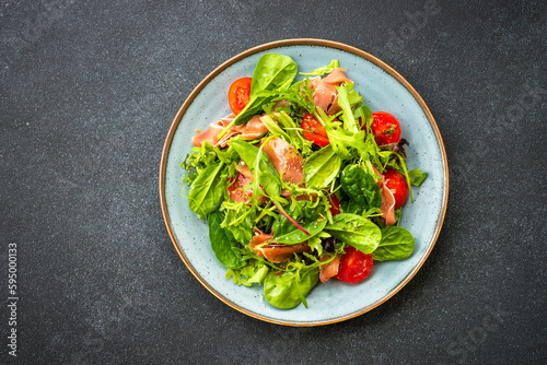 Green salad with fresh leaves, tomatoes and jamon at black table. Top view with copy space.