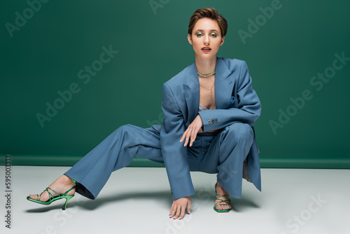 young woman with short hair sitting on haunches while posing in stylish pantsuit and heeled sandals on turquoise white background.