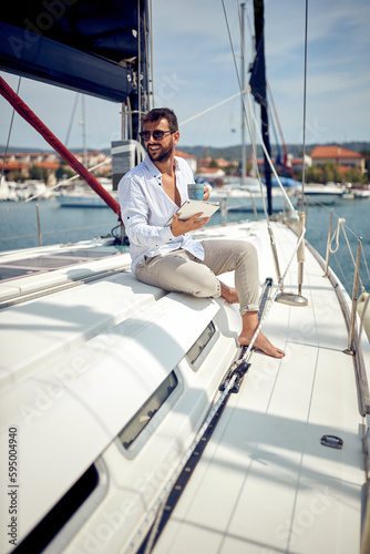 A young man with a tablet is sitting on the yacht and enjoying a coffee and beautiful scenery of the dock on the seaside. Summer, sea, vacation