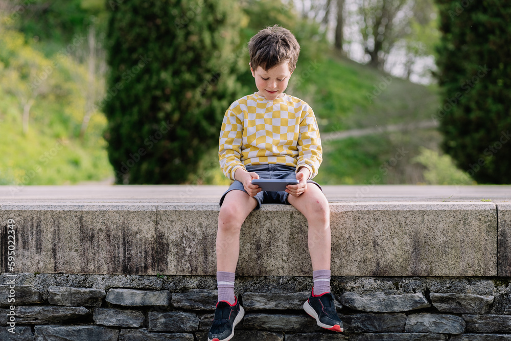 Smiling schoolboy sitting and looking at smartphone with digital devices in hands outdoors. Smiling schoolboy sitting and looking at smartphone with digital devices in hands outdoors.
