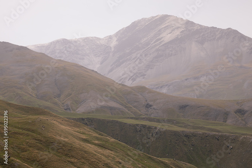 Majestic mountain landscape - mountain peak with glacier in soft light white pastel dim mist, yellow hilly folded slopes with dry alpine meadow in autumn, panorama view. Joyful landscape.