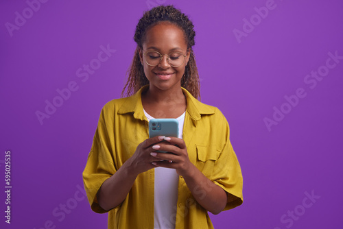 Young positive beautiful African American woman in casual clothes typing text on mobile phone using dating apps or instant messengers to find boyfriend stands on isolated lilac background