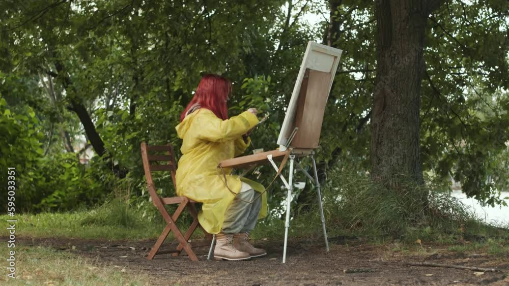Red haired girl in yellow raincoat sitting on wooden chair by canvas outdoors in park painting landscape Red haired girl in yellow raincoat sitting on wooden chair by canvas outdoors in park painting landscape