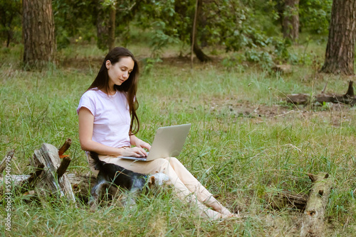 A beautiful young woman, girl with long brown hair sitting in a clearing in green forest with a little dog Yorkshire Terrier, working at laptop. Remote work, study. Student with a computer at nature.