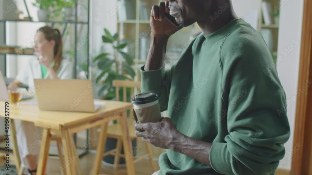 Cropped shot of black man in casual wear standing in open space office during workday, holding to go coffee cup and calling on mobile phone Cropped shot of black man in casual wear standing in open space office during workday, holding to go coffee cup and calling on mobile phone