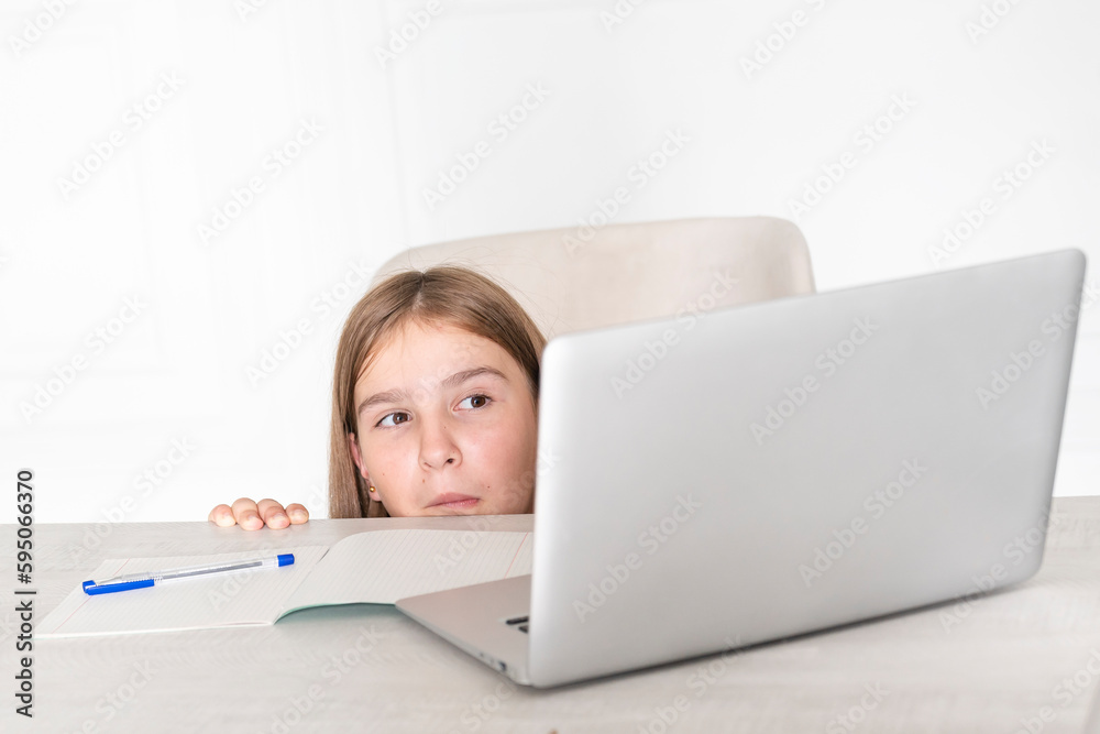 Bored and tired school girl hiding behind desk and looking at laptop. Studying online at home concept Bored and tired school girl hiding behind desk and looking at laptop. Studying online at home concept