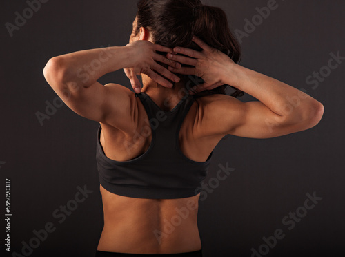 Sporty muscular woman with ponytail doing stretching workout of the shoulders, blades in sport bra, holding hands behind the head on dark grey background with empty space. Closeup portrait.