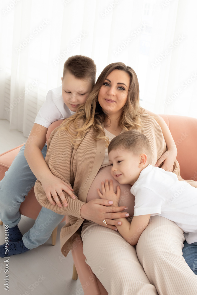 a cute little boy hugs his mother for a pregnant belly on a sofa in a large bright living room. smile of mom and brother in the background. waiting for replenishment in the family