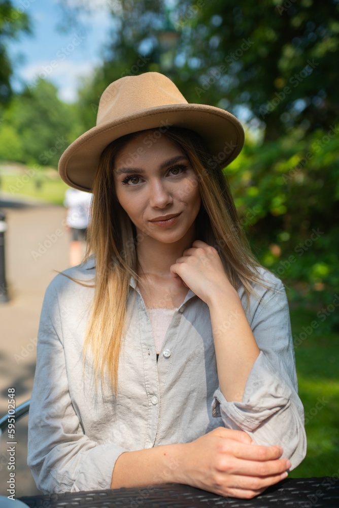 Young beautiful blonde woman in a wide-brimmed hat and pale gray shirt looking at the camera.