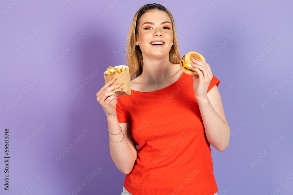 happy smiling pretty girl eats fast food, cheap burger, holding fries and burger. fast food and unhealthy diet. isolated on purple background. happy smiling pretty girl eats fast food, cheap burger, holding fries and burger. fast food and unhealthy diet. isolated on purple background.