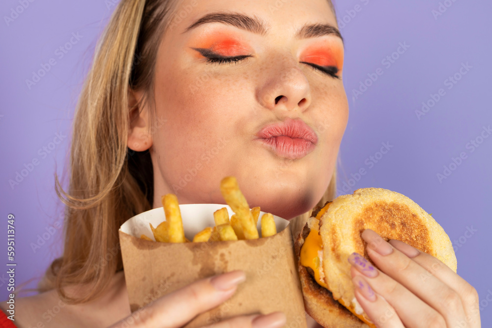 happy pretty girl eats fast food, cheap burger, holding fries and burger, closed eyes, dreams. fast food and unhealthy diet. isolated on purple background. happy pretty girl eats fast food, cheap burger, holding fries and burger, closed eyes, dreams. fast food and unhealthy diet. isolated on purple background.