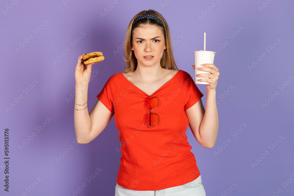 unhappy upset pretty girl eats fast food, cheap burger, holds a cheeseburger and soda. fast food and unhealthy diet. isolated on purple background. unhappy upset pretty girl eats fast food, cheap burger, holds a cheeseburger and soda. fast food and unhealthy diet. isolated on purple background.
