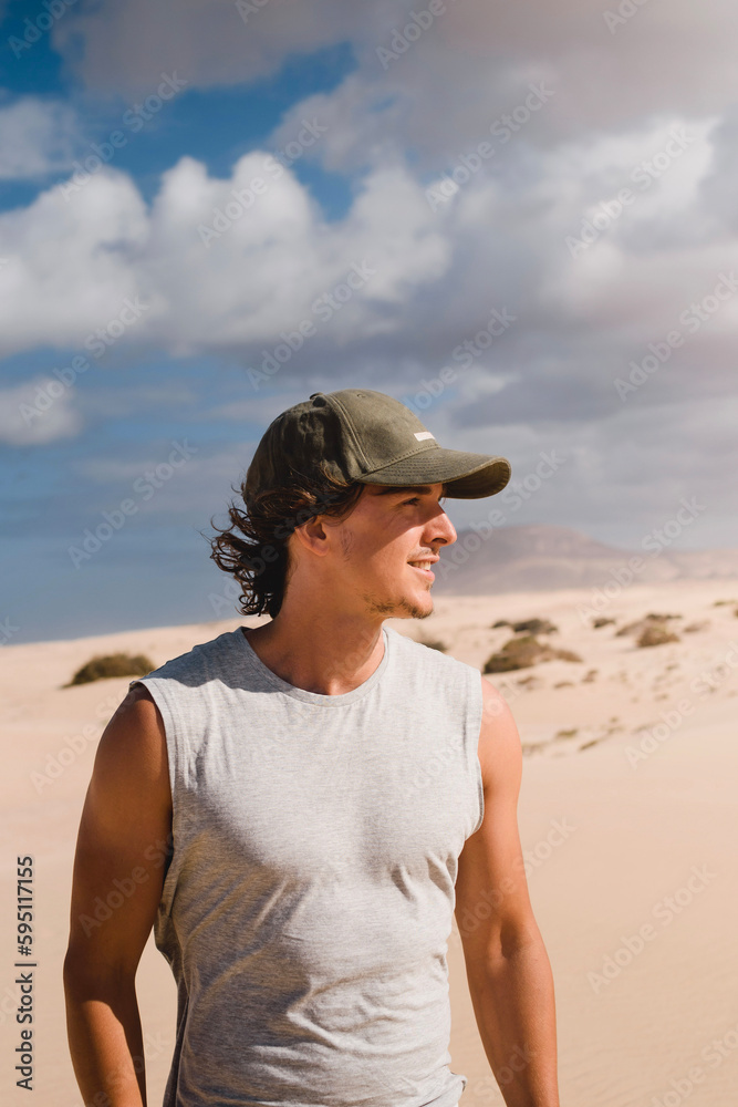 Portrait photo of smiling young man with cap in the desert of the dunes of Corralejo in Fuerteventura, looking at the horizon, in summer, during a vacation trip through the Canary Islands.
