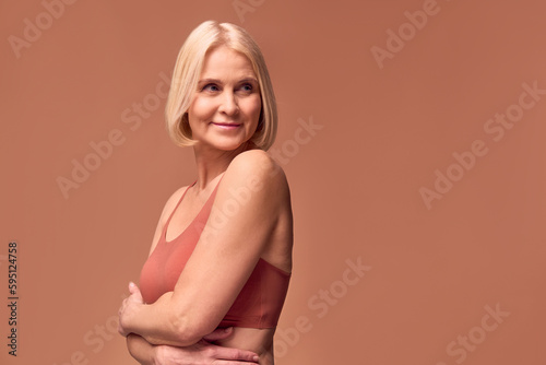 Attractive, beautiful, athletic older woman in underwear standing near a beige background with her arms crossed and looking away. Body care concept.