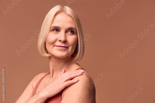 Portrait of a beautiful older woman with blond hair looking to the side on a beige background and holding a hand on her shoulder.