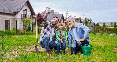 Cheerful man taking selfie photo with smartphone of grandfather, father, son and grandson sitting in garden and smiling to camera. Planting trees concept. Making picture of three generations.