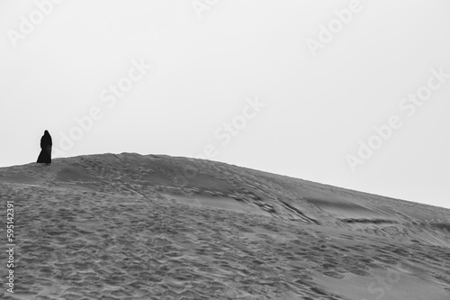 The burqa's mystery: alone muslim woman with black burqa in the vast desert under pale sky (in black and white)