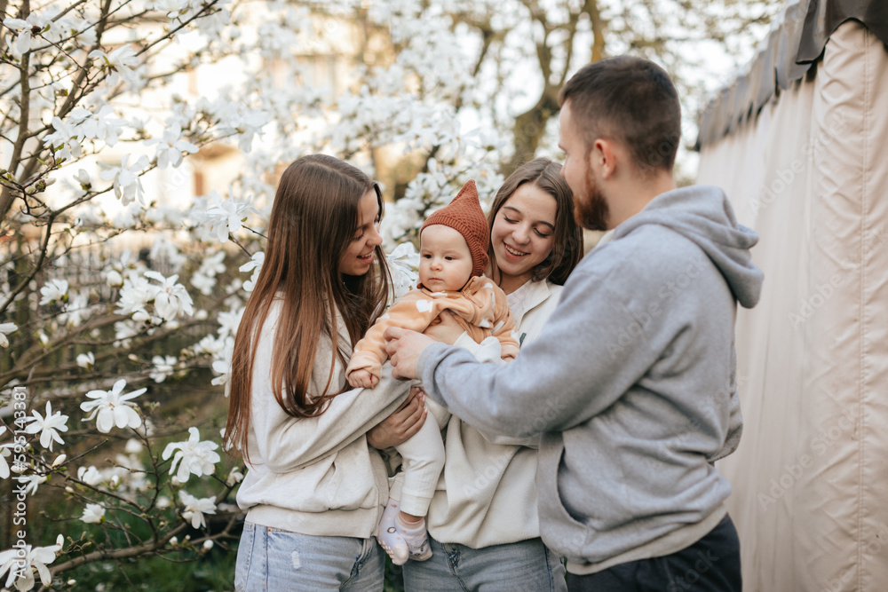 Fototapeta premium A little baby in the arms of twin sisters was delighted when she saw her father in the blooming garden. Lifestyle. Family concept. Father and Daughter. 