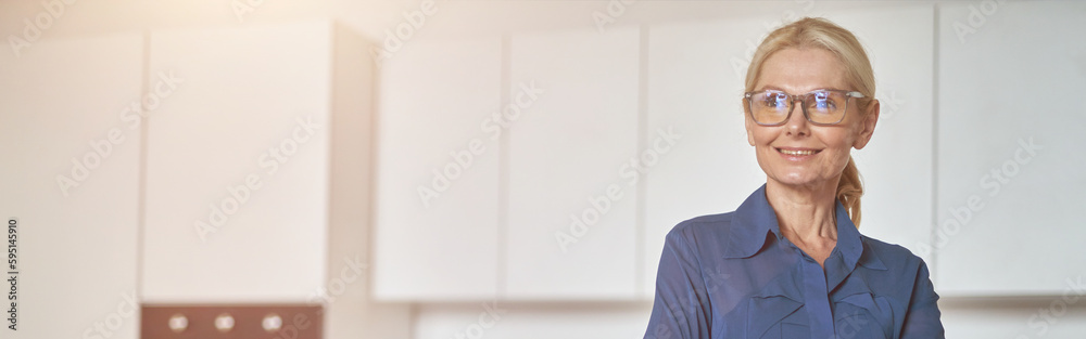 Confident mature businesswoman in glasses smiling aside, working at home using laptop, standing with arms crossed in modern kitchen