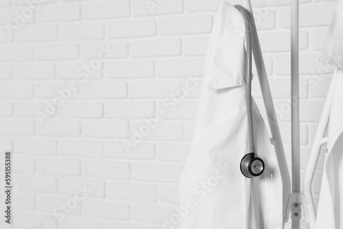 Medical uniforms and stethoscope hanging on rack near white brick wall. Space for text photo