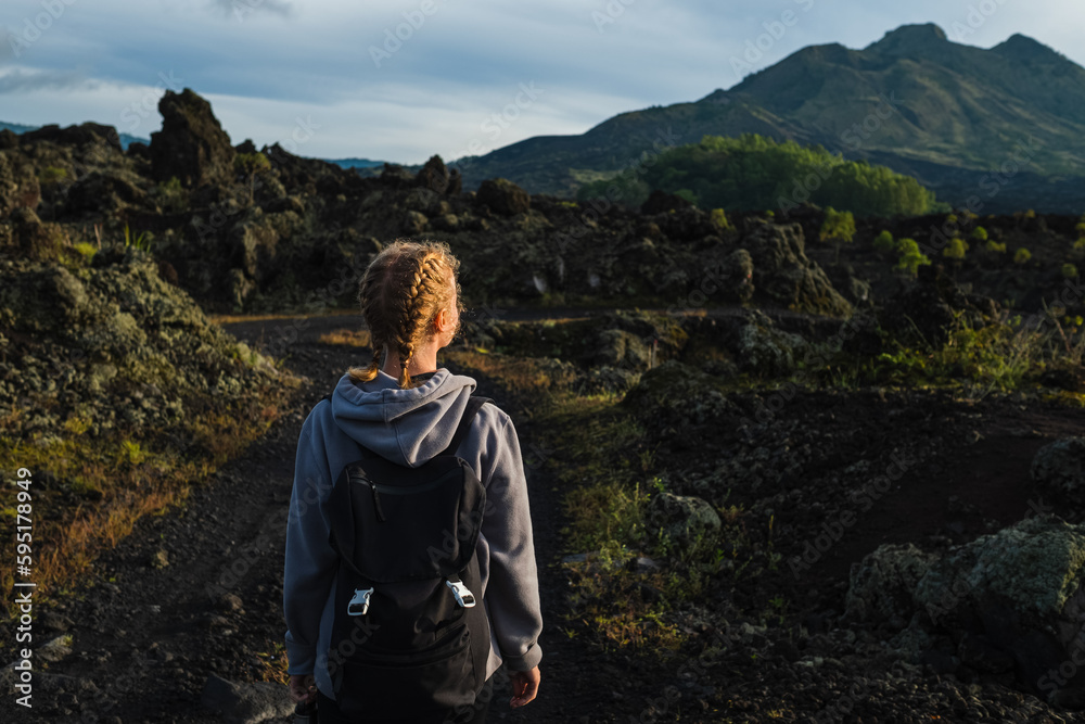 A young girl traveler with a backpack walks in the mountains against the backdrop of the Batur volcano on the island of Bali in Indonesia. A young girl traveler with a backpack walks in the mountains against the backdrop of the Batur volcano on the island of Bali in Indonesia.