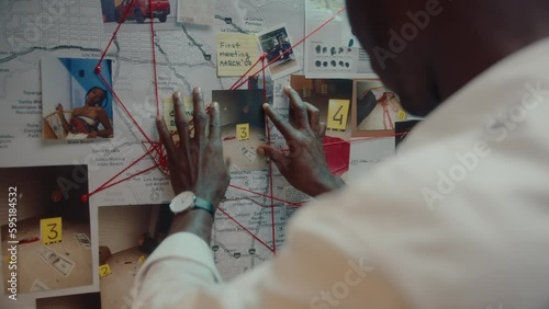 Black detective attaching crime scene photo to pinboard on the wall in his office, looking at clues connected with red thread, working on murder investigation photo