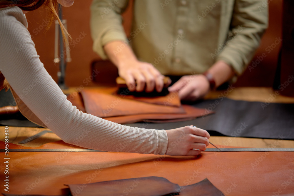 Obraz premium woman's arms measuring leather for sewing, man using brush to clean the surface of material from the dust. close up cropped side view shot, focus on arms. blurred background