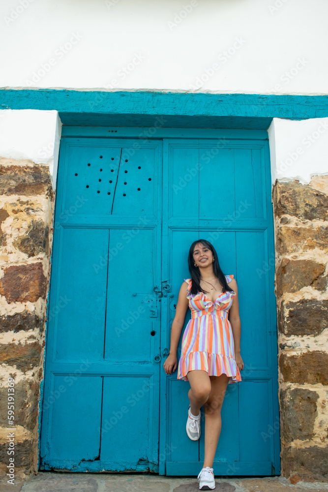 Beautiful young Colombian Latina smiling wearing a colorful dress. In the background an old colonial house with a blue painted wooden door and stone walls.